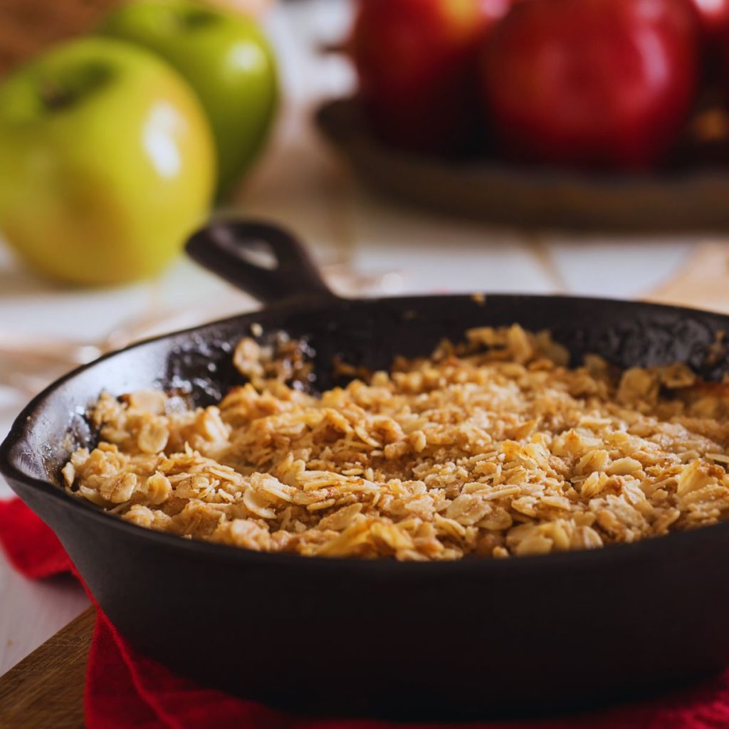 an apple crisp in a cast iron skillet on a red napkin with apple in the background and a flaky oat topping